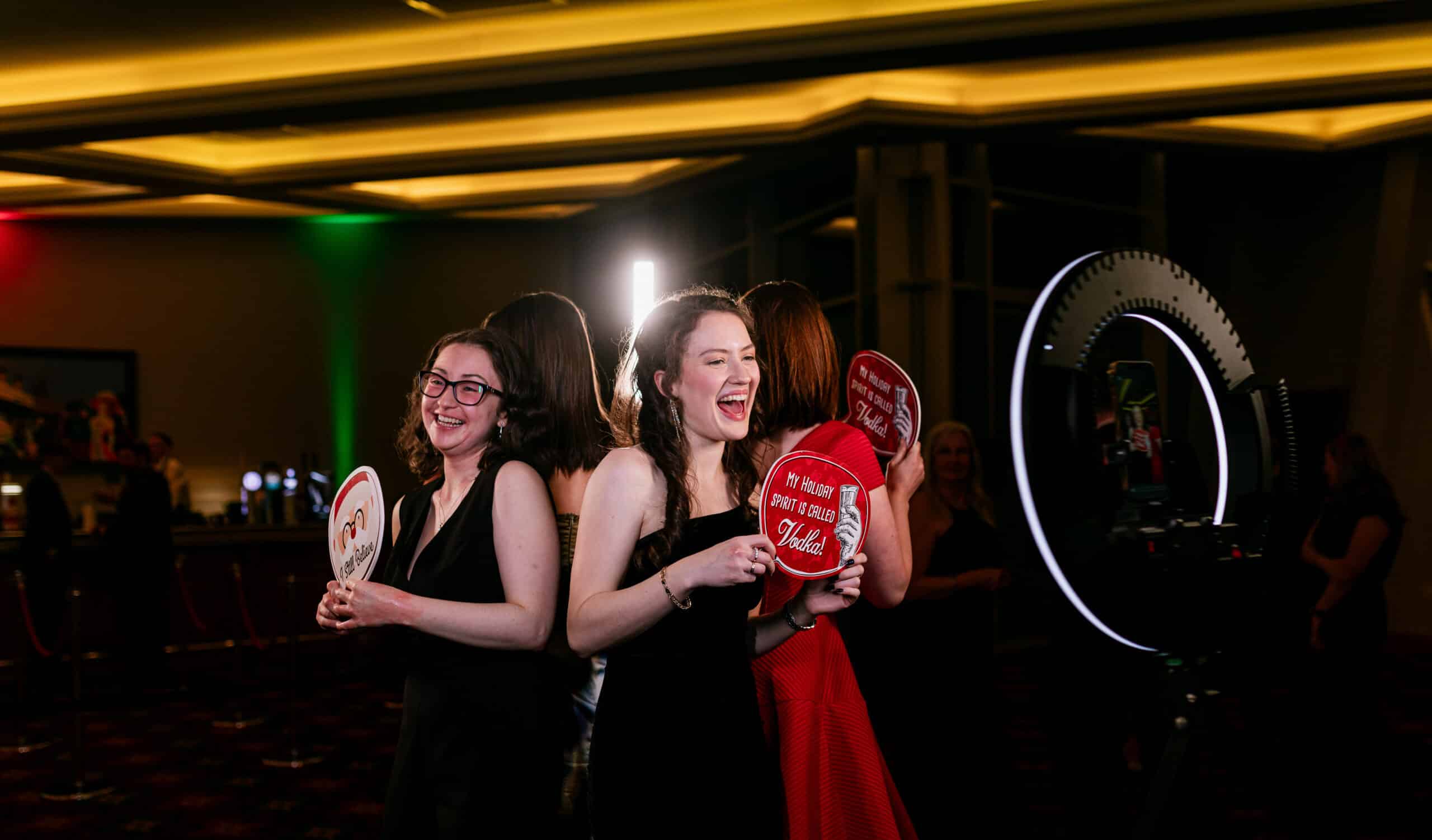 Ladies and gentlemen at a party joyfully posing with signs in front of a photo booth, capturing fun moments.