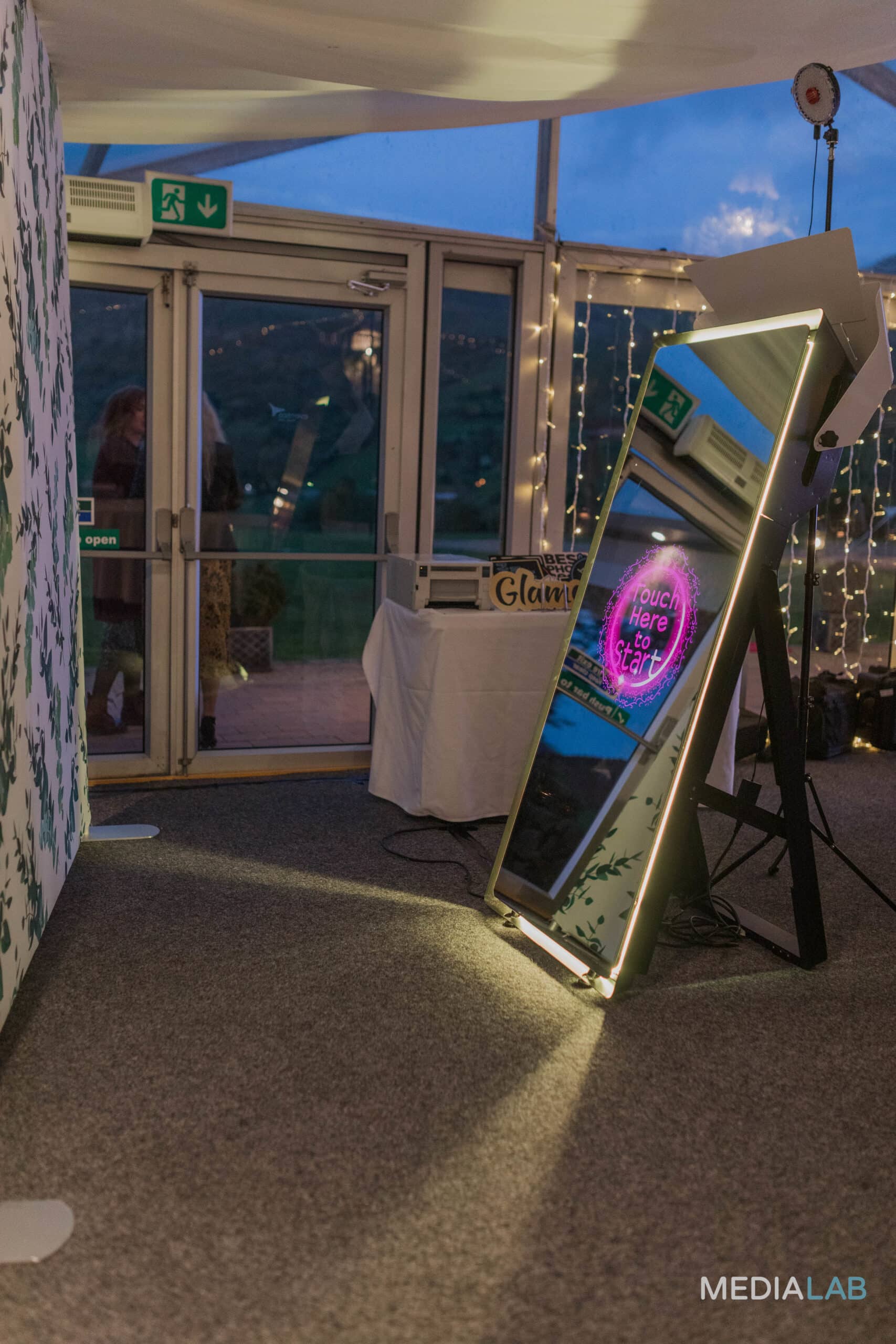 Mirror photo booth setup with illuminated frame, displaying "Touch Here to Start," in a decorated event space, ready for guests at a wedding or party.