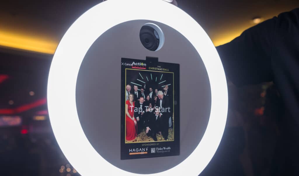 Corporate photo booth setup featuring a ring light and touchscreen display with "Tap to Start" prompt, promoting engagement at events like the Christmas Ball.