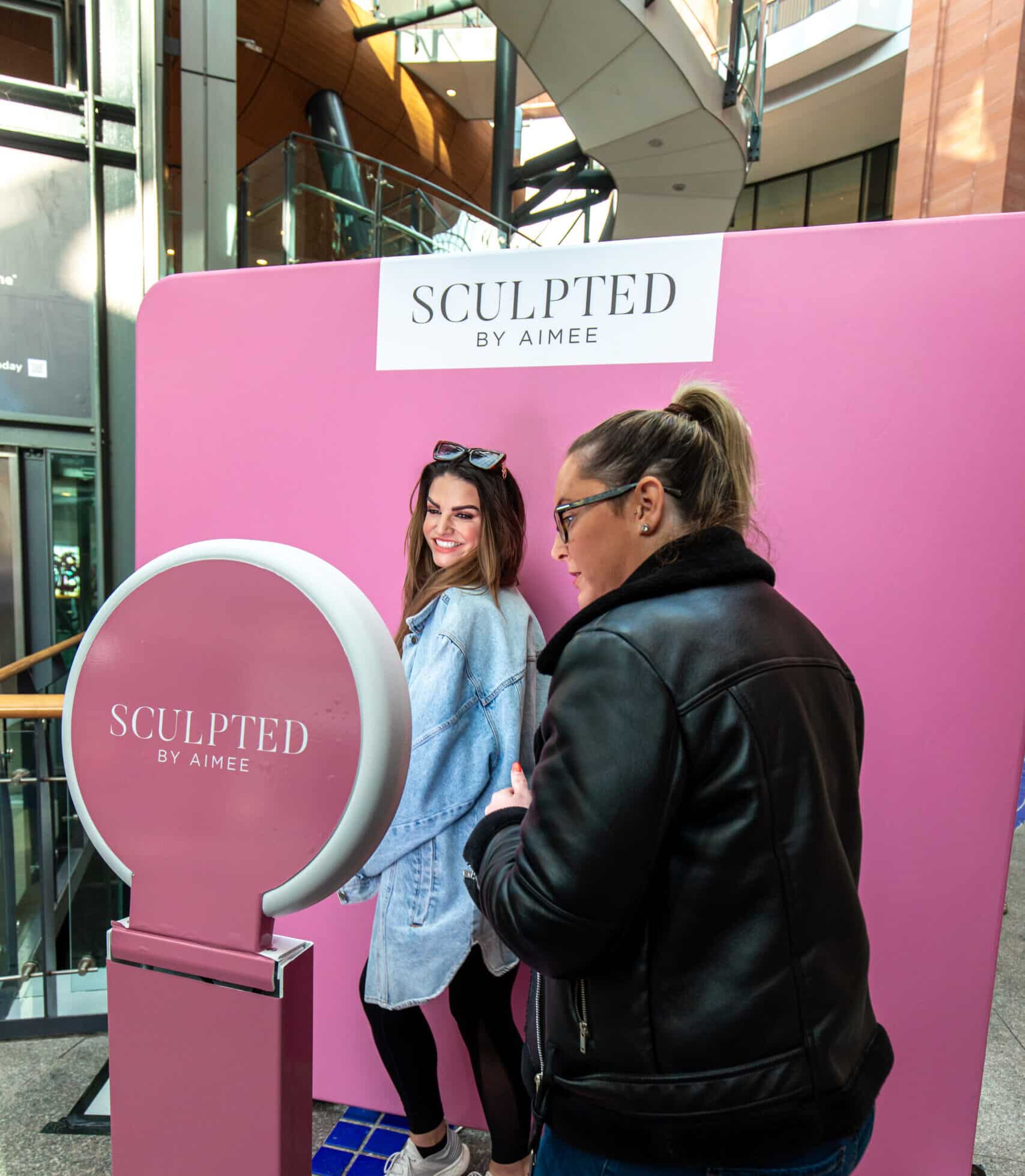 Photo booth setup featuring a pink backdrop with "SCULPTED BY AIMEE" signage, two women interacting, one posing playfully, in a vibrant event space.