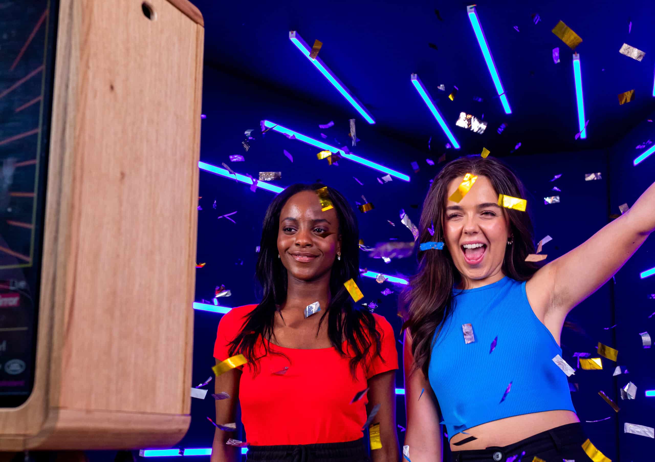 Two women posing for a photo with an open air photo booth in front of a neon backdrop